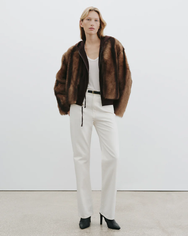 Woman wearing a brown fur coat, white top, and white pants against a white background