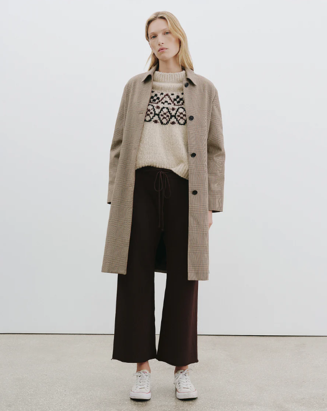 Woman wearing a beige coat, patterned sweater, and dark pants on a white background