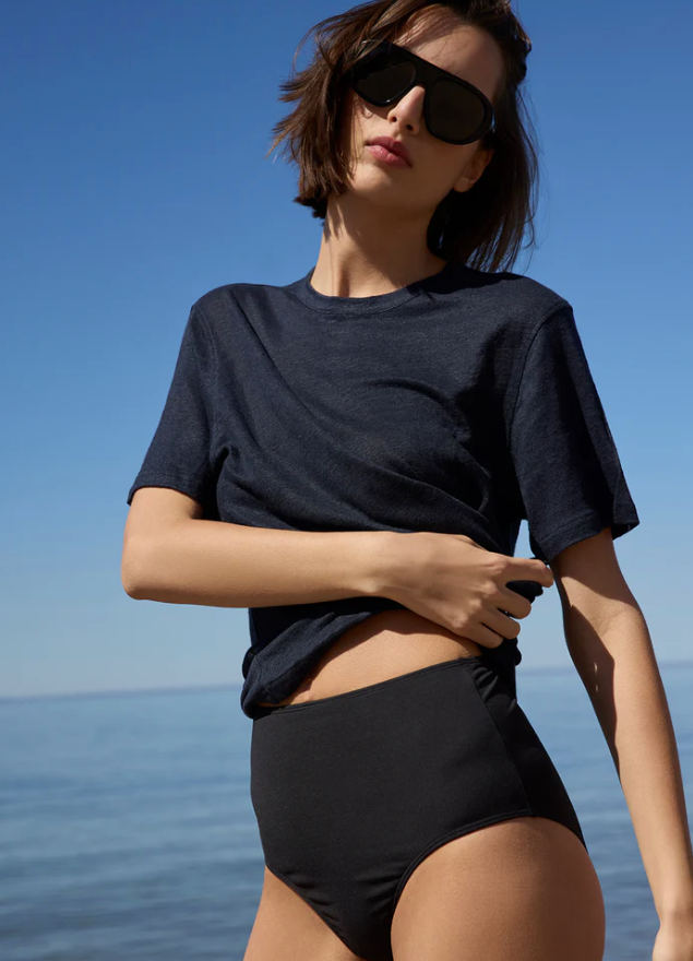 A woman wearing a dark navy blue short-sleeve crewneck t-shirt and black high-waisted bikini bottoms, standing outdoors against a clear blue sky and sea background.