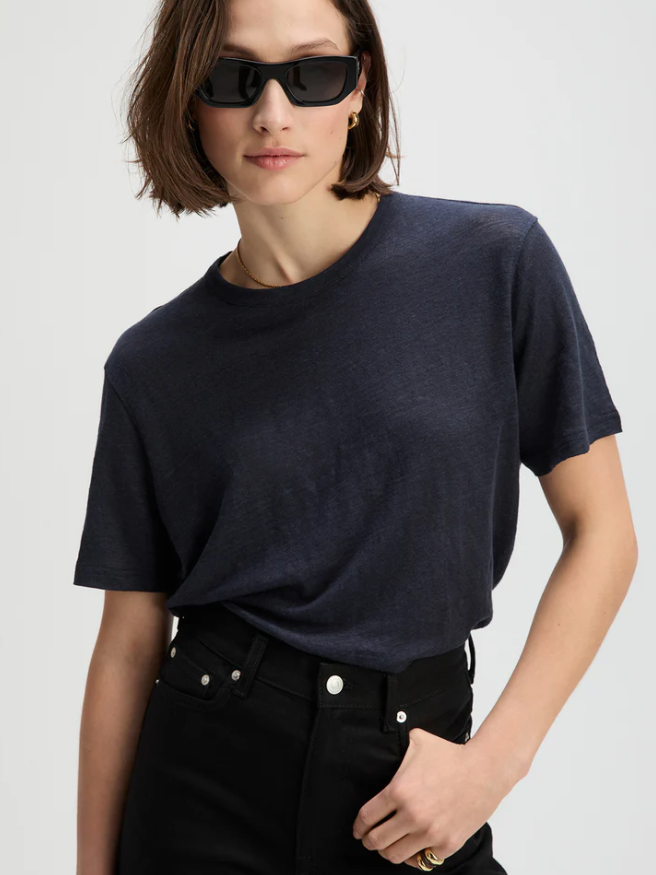 A woman wearing a dark navy blue short-sleeve crewneck t-shirt and black jeans, styled with black sunglasses against a light gray background.