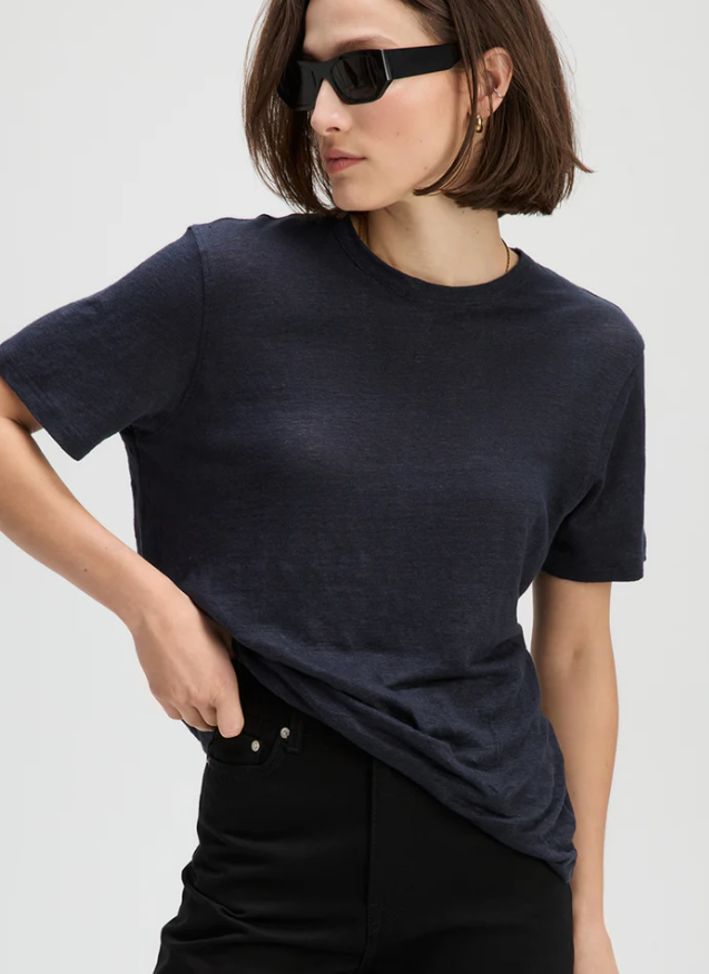 A woman wearing a dark navy blue short-sleeve crewneck t-shirt and black pants, styled with black sunglasses against a light gray background.