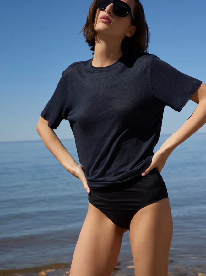 A woman wearing a dark navy blue short-sleeve crewneck t-shirt and black swim briefs, standing outdoors by the ocean with a clear blue sky in the background.