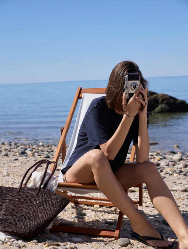 A woman sits on a wooden beach lounge chair on a pebble shore, holding a vintage camera to her face. She is wearing a dark navy t-shirt and black flip-flops, with a dark woven tote bag beside her and the blue sea and sky in the background.