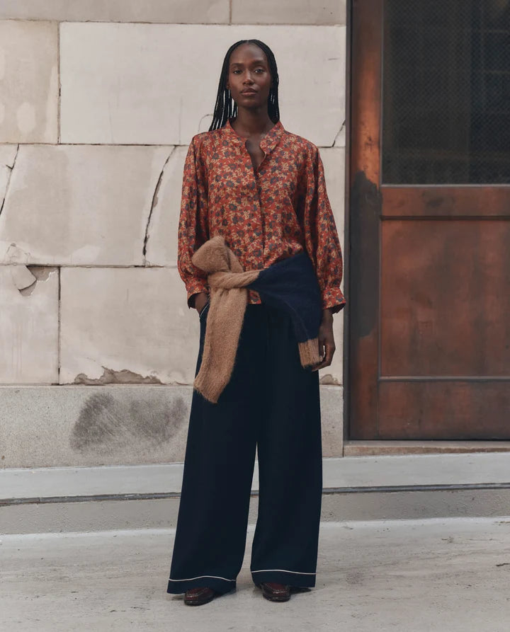 A woman stands outdoors in front of a stone wall and wooden door, wearing a rust-orange floral long-sleeve button-up shirt, dark navy wide-leg trousers, and a tan cardigan tied around her waist.