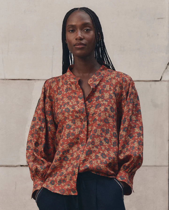 A woman with long black braids wearing a rust-orange long-sleeve button-up blouse with a dense multicolor floral print, paired with dark trousers, standing against a light beige stone wall.