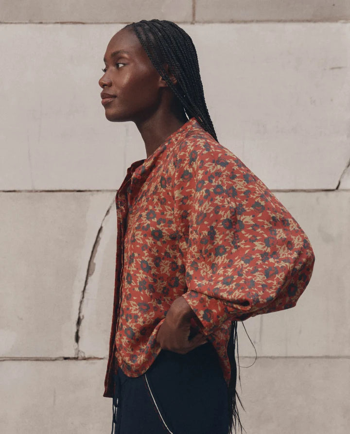 A woman in profile wearing a loose, long-sleeve button-up shirt with a rust red and navy floral pattern, paired with dark navy bottoms, standing against a light beige stone wall.