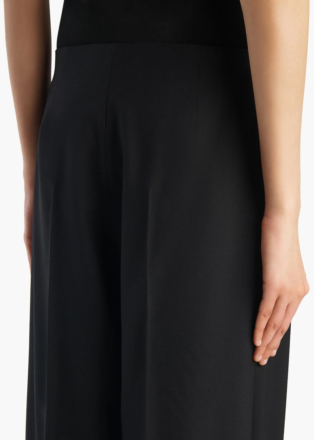Close-up of a model wearing black wide-leg trousers, showing the side profile, hand, and part of the torso against a plain white background.