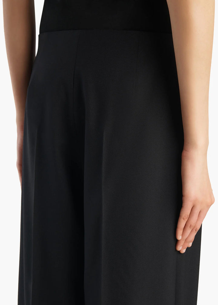 Close-up of a model wearing black wide-leg trousers, showing the side profile, hand, and part of the torso against a plain white background.
