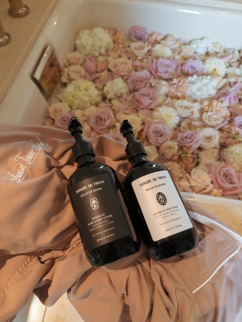 Two bottles of Sanfte de Fruta products on a towel with a bathtub filled with flowers in the background.