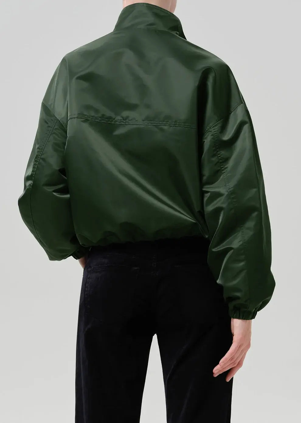 A person shown from the back wearing a dark green shiny jacket and black pants against a light gray studio background.