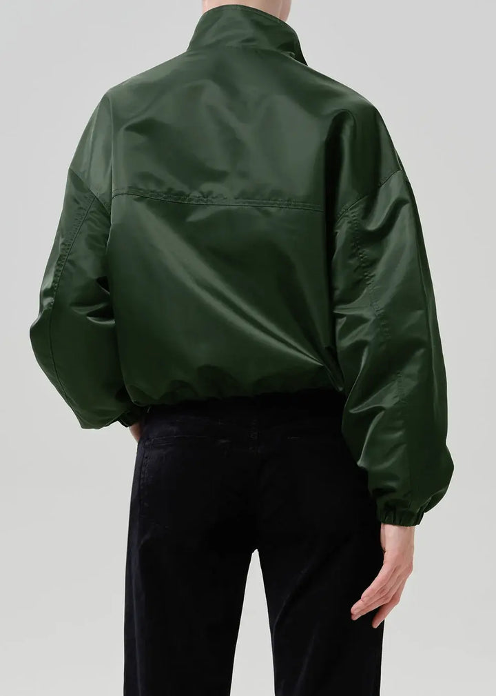 A person shown from the back wearing a dark green shiny jacket and black pants against a light gray studio background.