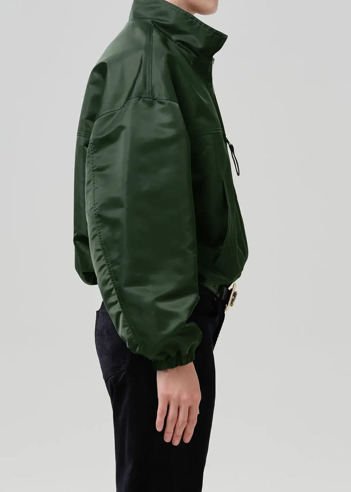 A side profile view of a person wearing a dark forest green lightweight jacket with a high collar and black pants, set against a solid light gray background.