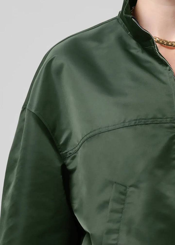 Close-up of a person wearing an olive green nylon jacket with a high collar and a gold chain necklace, against a light gray background.