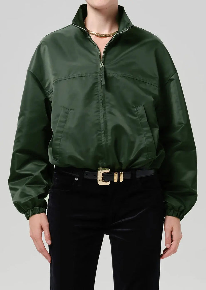 A person wearing a dark green satin zip-front jacket with a stand collar, paired with black pants and a black belt with a gold buckle, against a light gray studio background.