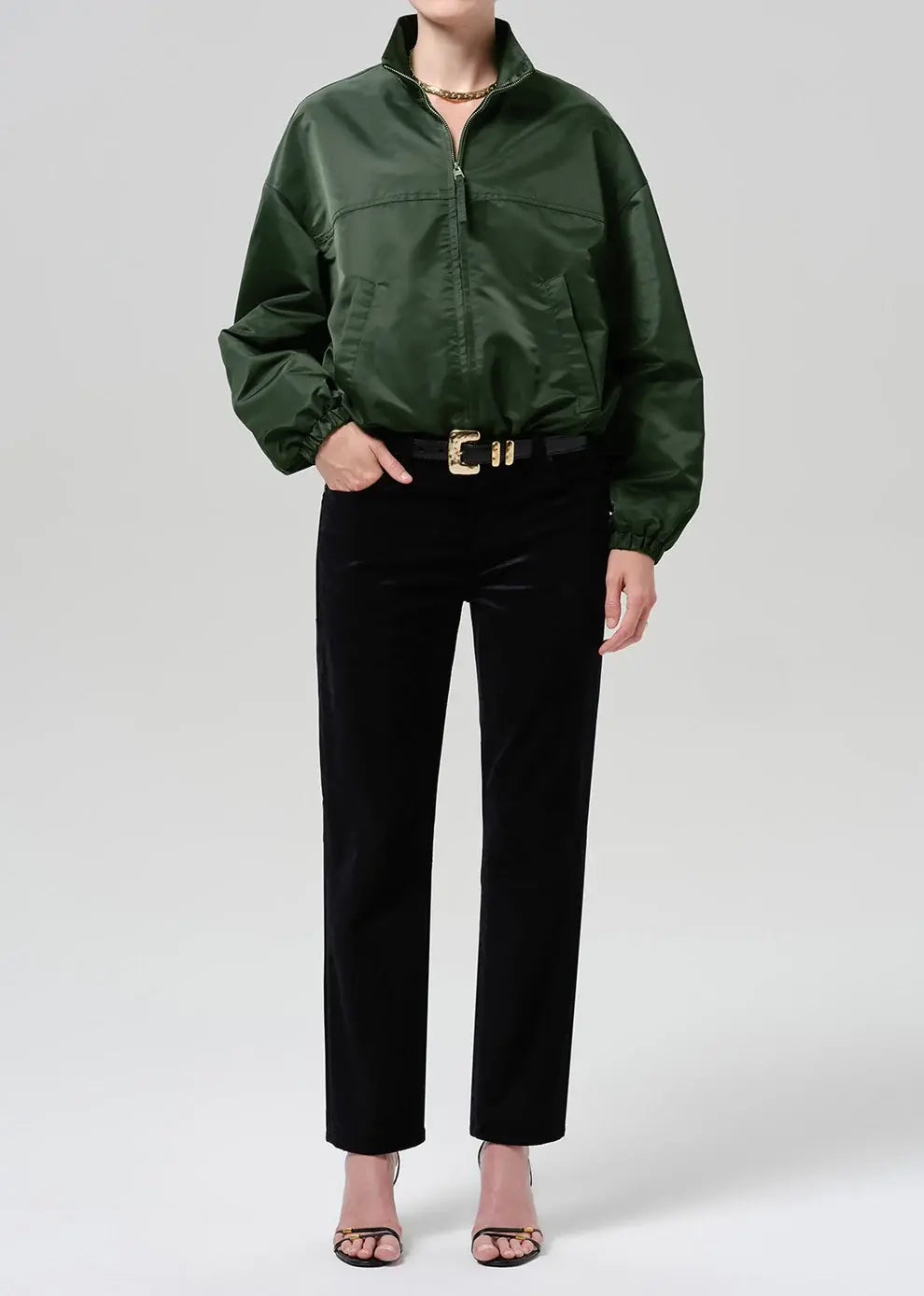 A model wearing a black satin bomber jacket, black velvet trousers, and a black belt with a gold buckle, standing against a light gray studio background.