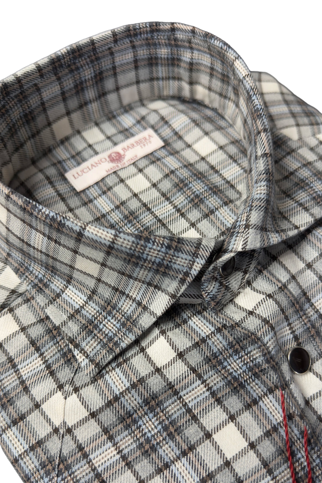 Close-up of a plaid button-down shirt collar and upper chest area, showing a white neck label with red text and a black button. The fabric has a gray, black, and white plaid pattern.