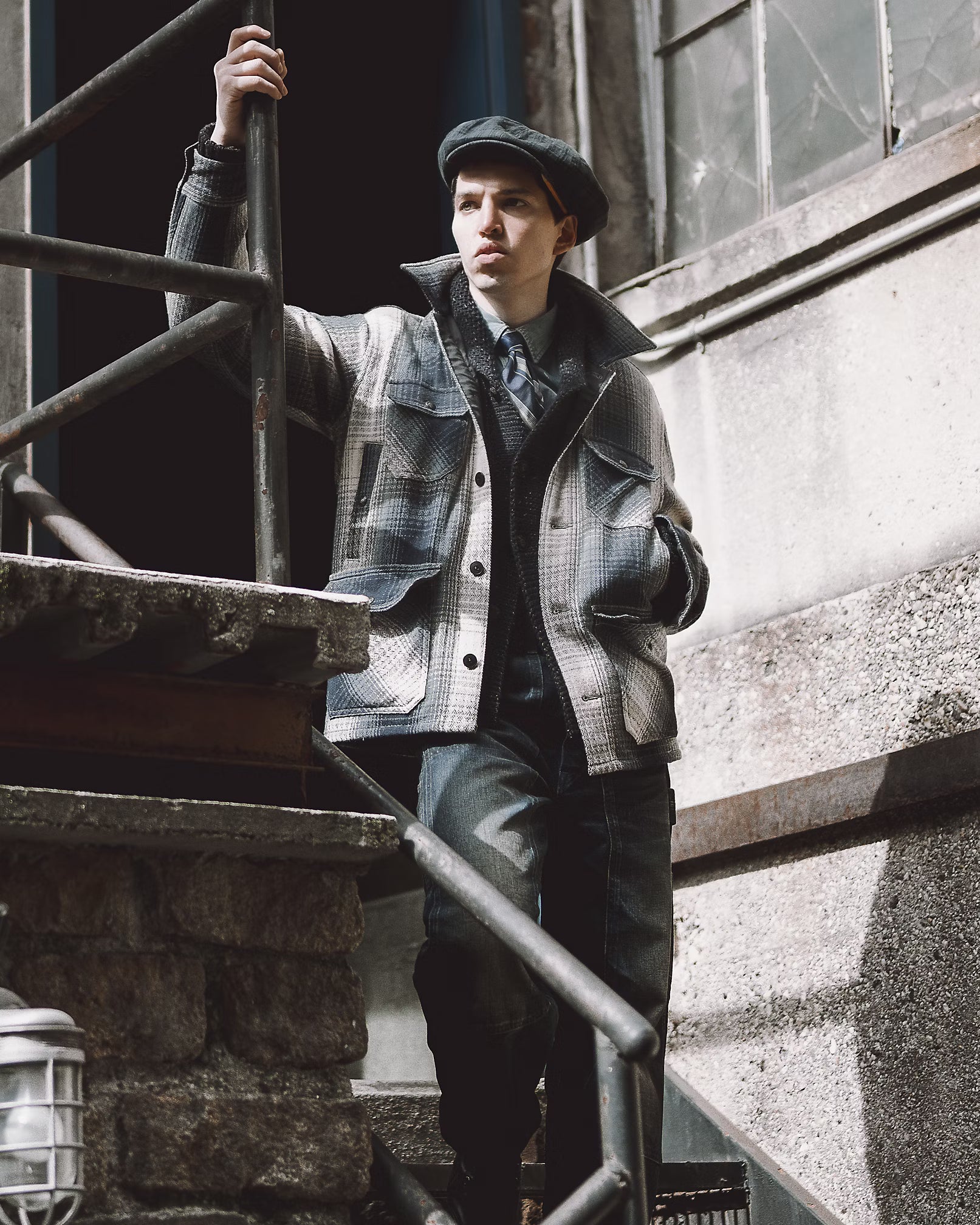 A man wearing a vintage-style plaid jacket, dark trousers, and a flat cap stands on outdoor stone stairs next to a building.