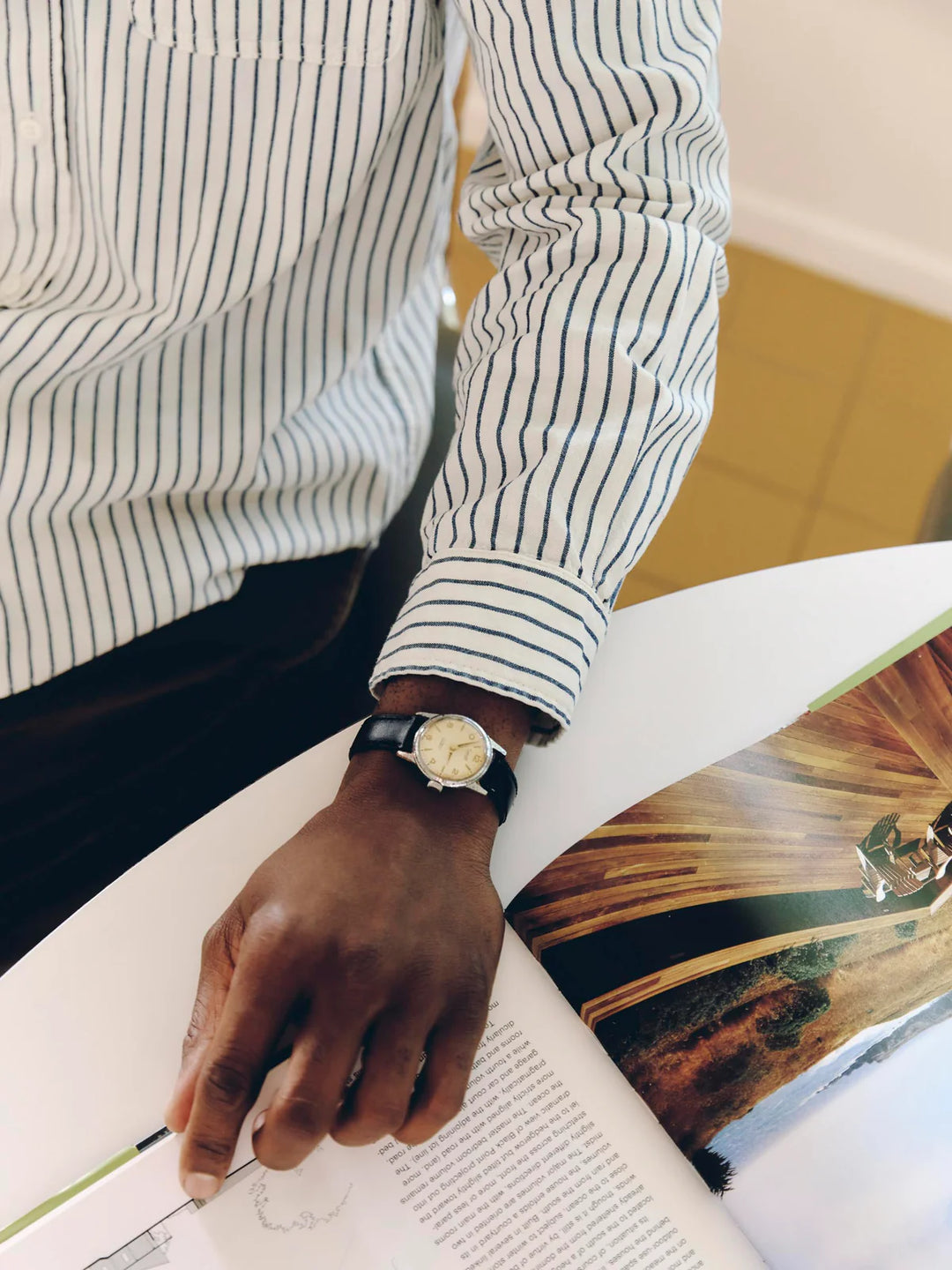 Person wearing a striped shirt and watch, holding an open book with a scenic cover.