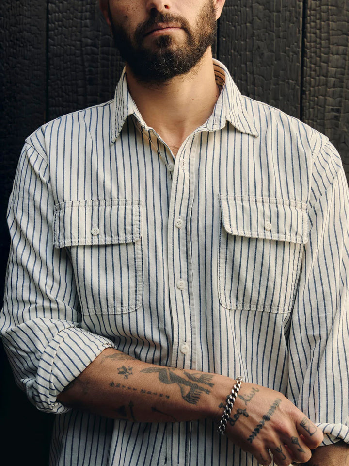 Man wearing a striped shirt with tattoos on his arms against a dark textured wall.