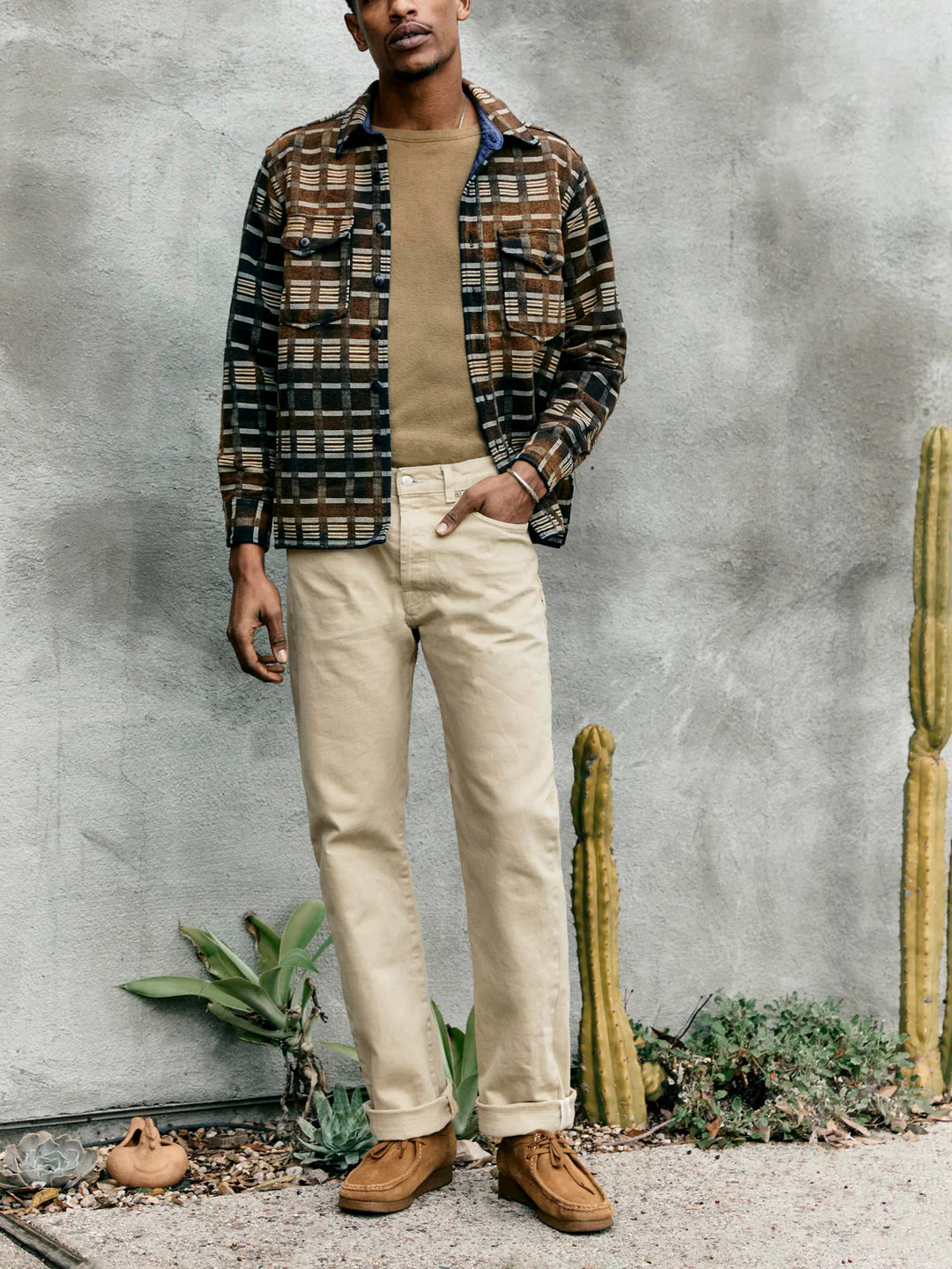 Man wearing a patterned shirt, beige sweater, and pants standing against a textured wall with cacti.
