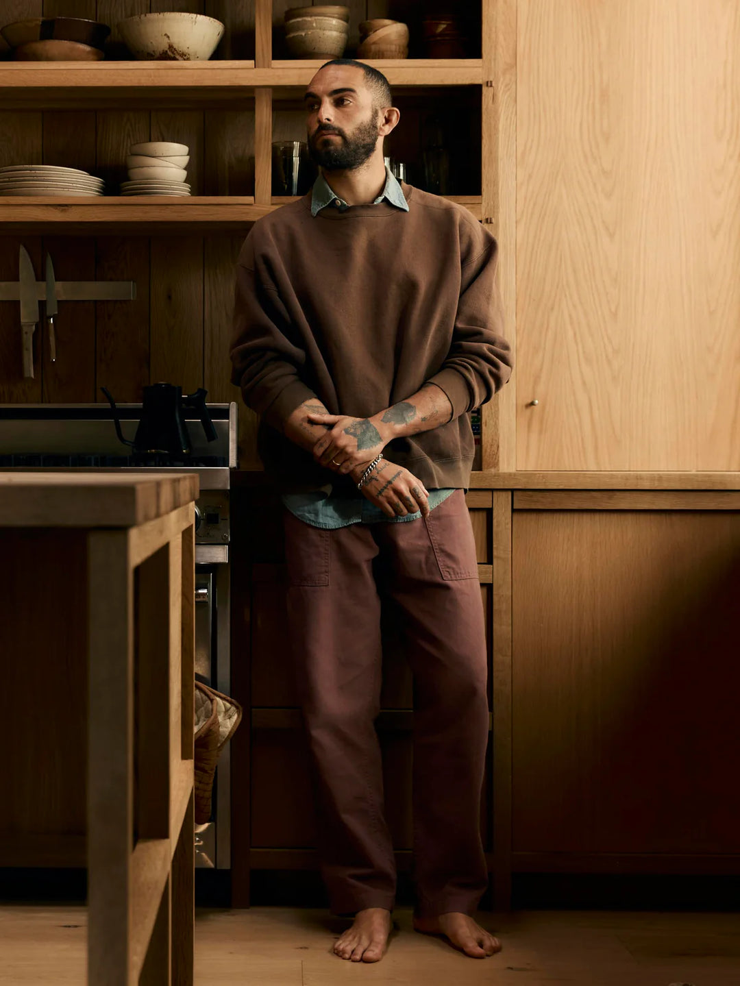 Man standing in a wooden kitchen wearing a brown sweater and dark pants.