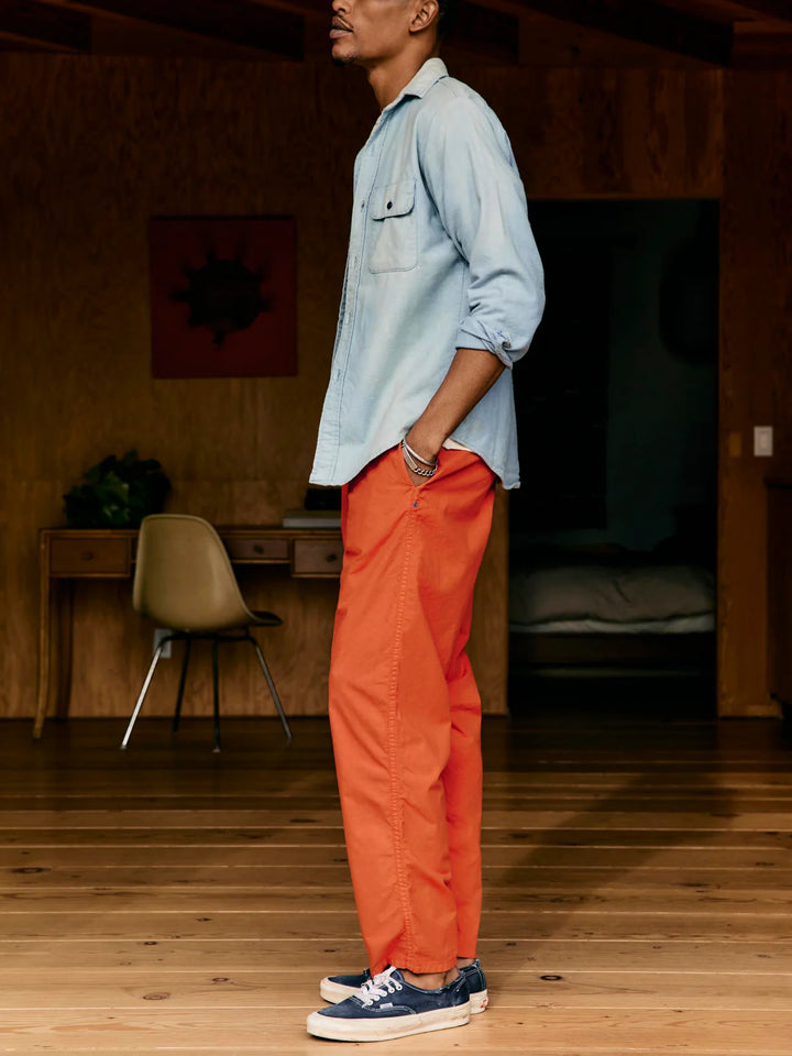 Person wearing a light blue shirt and orange pants standing in a room with wooden floor and furniture.
