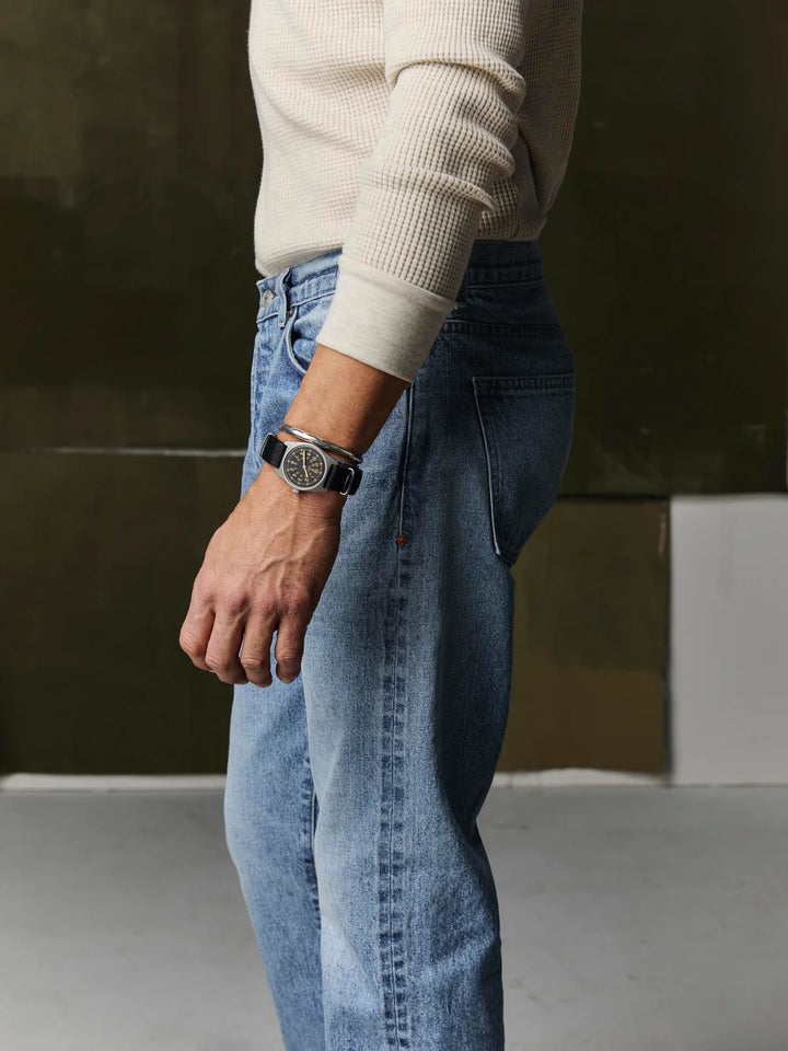 Person wearing a light-colored sweater and blue jeans with a watch on a neutral background