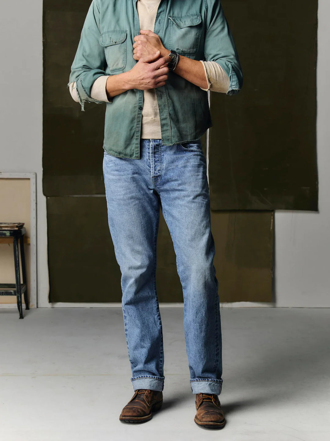Person wearing a green jacket, beige shirt, blue jeans, and brown shoes in an indoor setting.