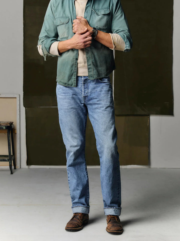 Person wearing a green jacket, beige shirt, blue jeans, and brown shoes in an indoor setting.